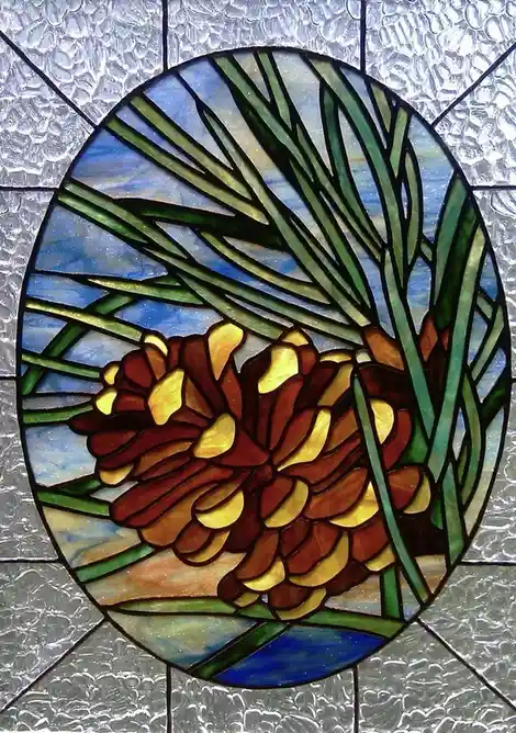 A stained glass window panel depicts a detailed pine cone surrounded by green pine needles against a blue sky background…