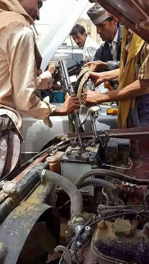 Three men are gathered around an open car hood, with one man holding a rifle and another holding a wrench, appearing to work…