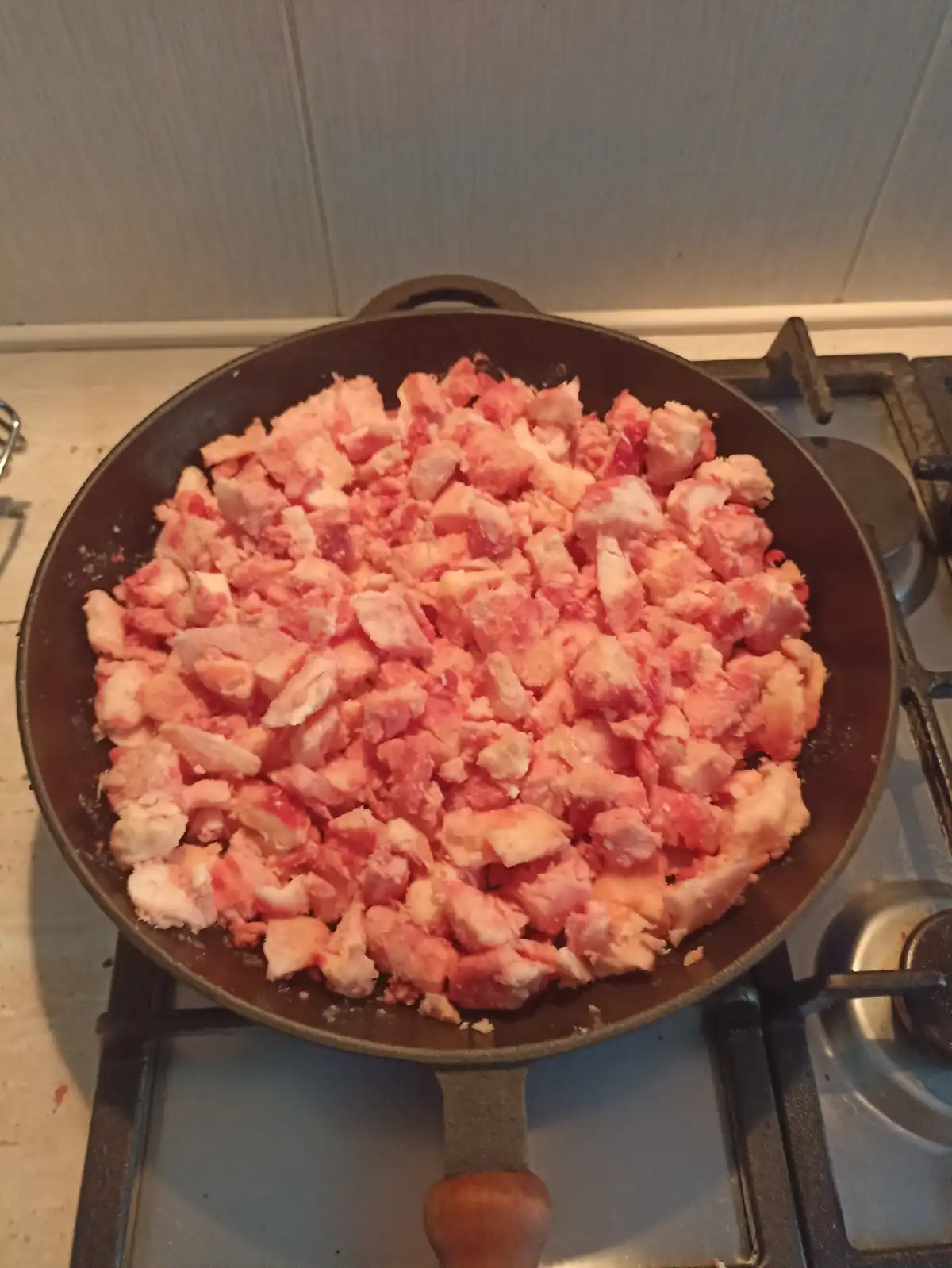 A black cast iron skillet sits on a gas stove burner, filled with a large quantity of chopped raw bacon pieces. The bacon is…