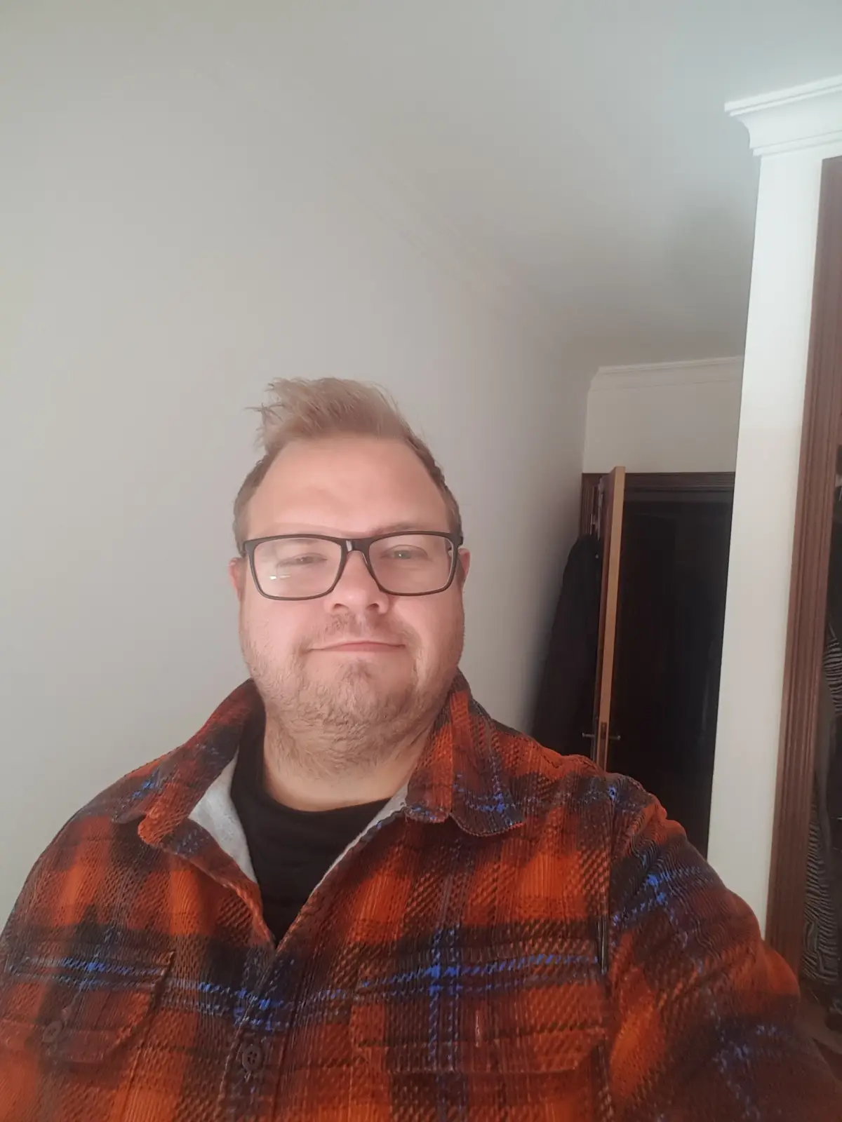 A man with glasses and a beard, wearing an orange and blue plaid flannel shirt over a black t-shirt, smiles smugly at the…