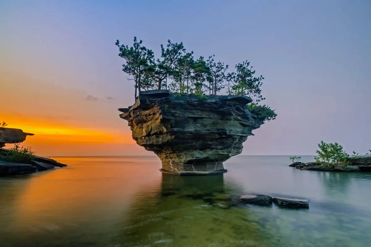 A serene landscape photo shows a large, flat-topped rock formation rising from calm water at sunset, with several pine trees…