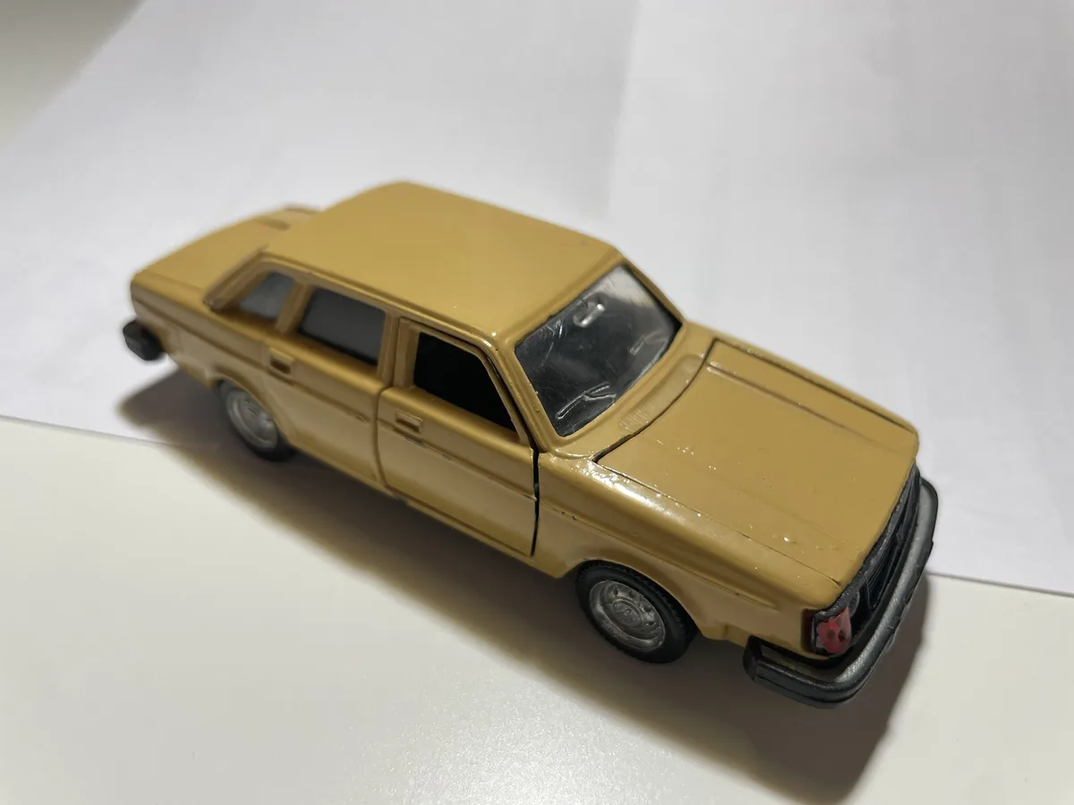 A mustard-yellow toy car, likely a Volvo 240, is photographed on a white surface. The car appears worn, with visible scuffs…