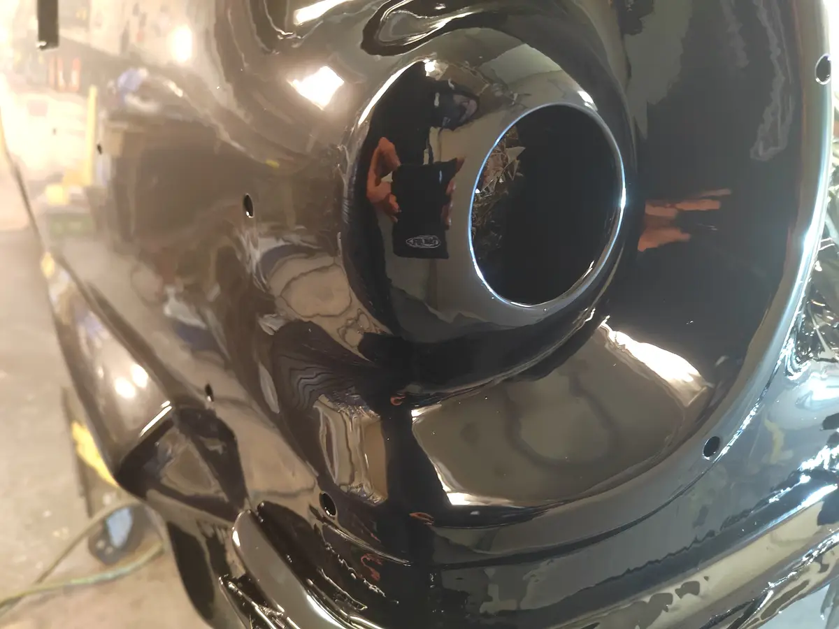 A close-up of a highly polished chrome exhaust tip reflects the person holding a phone with a 'DRL' logo, humorously…