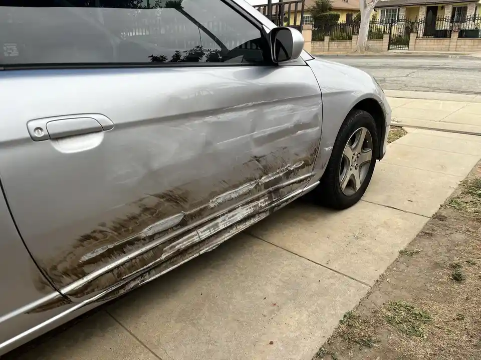 A silver car is parked on a concrete driveway with visible dirt and grime streaks along its lower side panel, contrasting…
