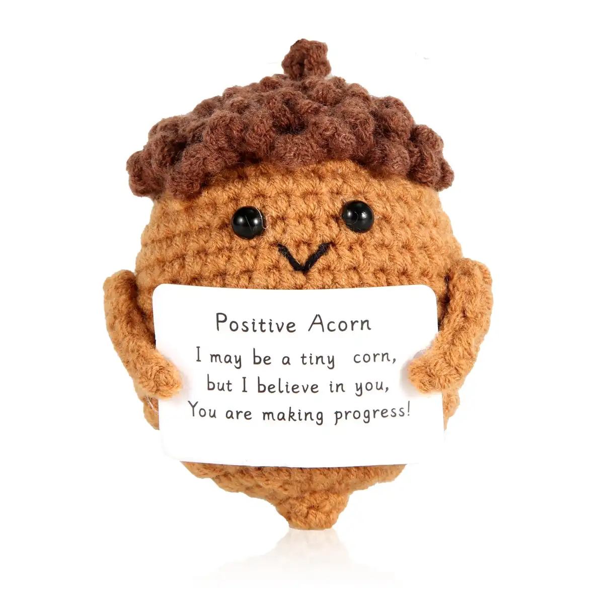 A handmade, crocheted acorn plush toy with a smiling face and black button eyes holds a white sign. The sign reads 'Positive…