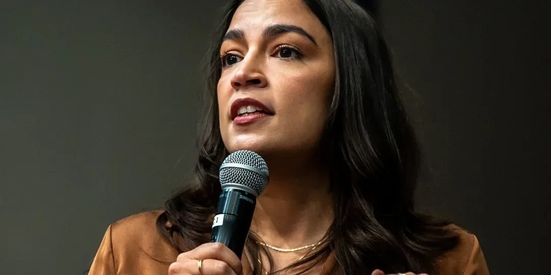 a woman with dark hair and a brown top holds a Shure microphone while speaking, her expression showing mild nervousness. The…