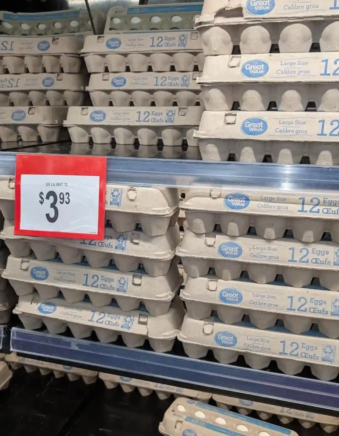 A supermarket shelf displays stacks of Great Value brand egg cartons labeled 'Large Size 12 Eggs'. A red-bordered price tag…