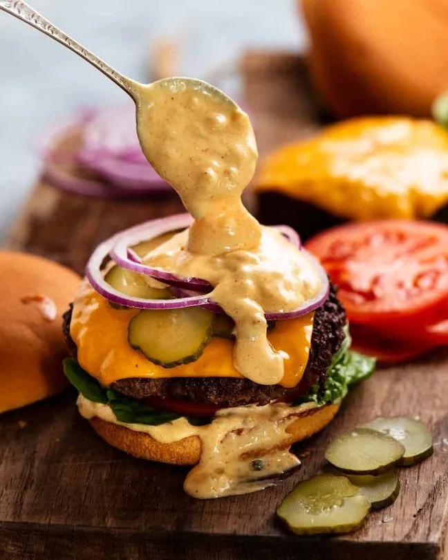 a close-up photo of a burger being generously drizzled with a thick, creamy sauce from a spoon. The burger has a beef patty…