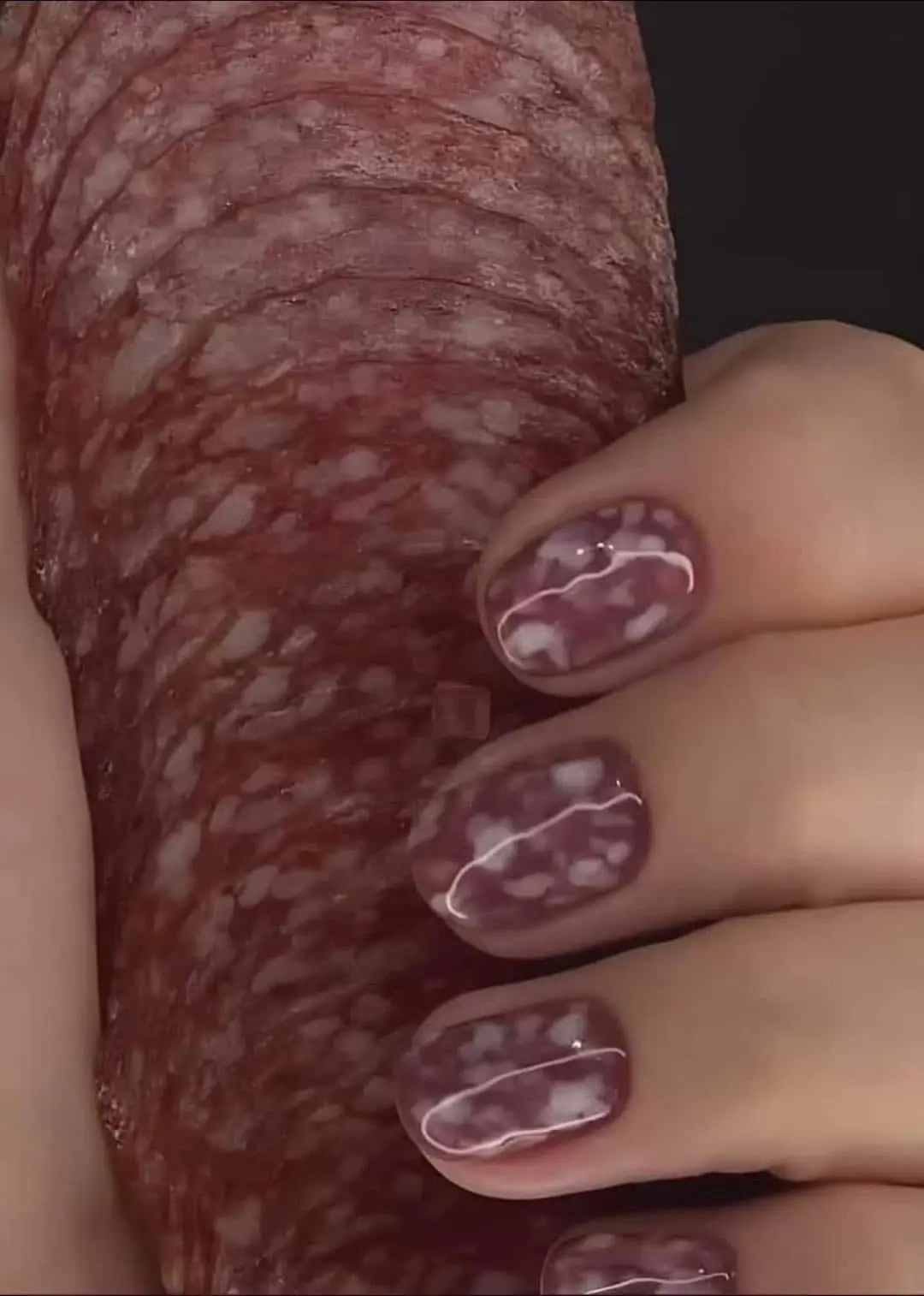 A close-up image shows a hand with fingernails painted to perfectly mimic the marbled red-and-white pattern of salami, held…