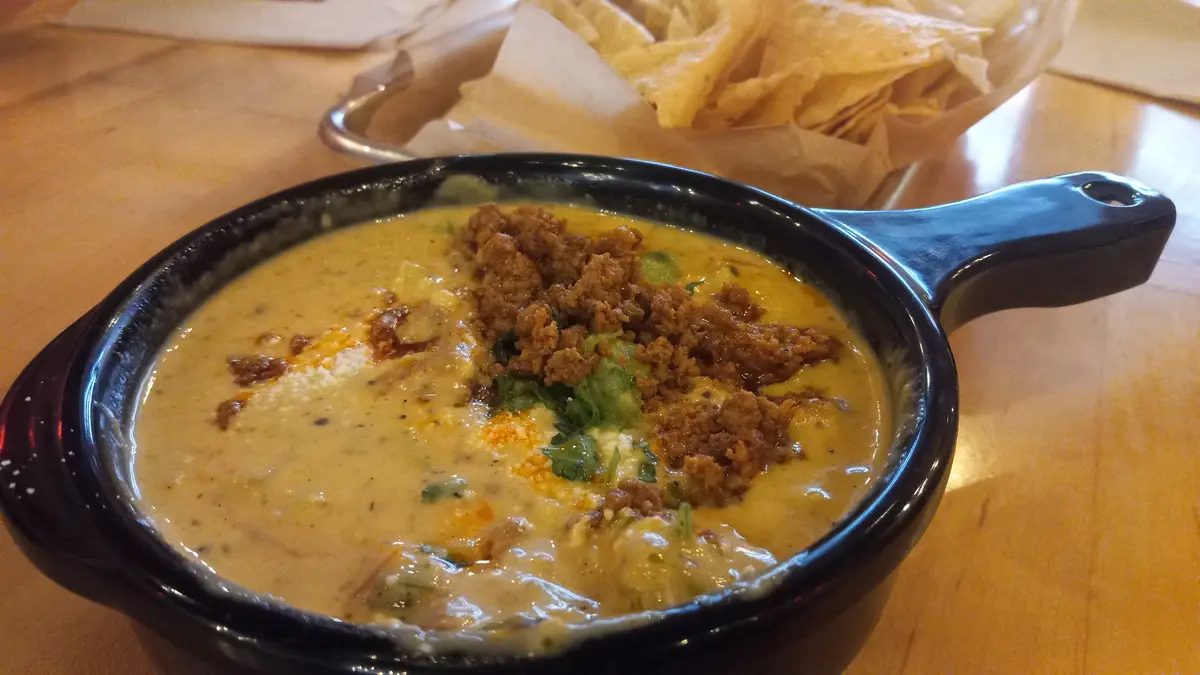 A close-up photo shows a black ceramic bowl filled with creamy yellow cheese dip, topped with brown crumbled meat, green…