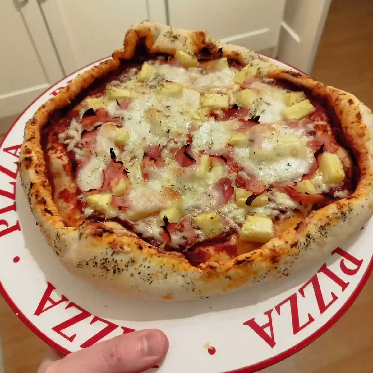 A close-up photo shows a freshly baked Hawaiian pizza on a white plate with red 'PIZZA' lettering around the rim, held by a…