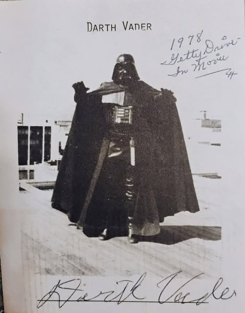 A black-and-white photo of Darth Vader standing in a snowy landscape, wearing his iconic helmet and cape. The image has…