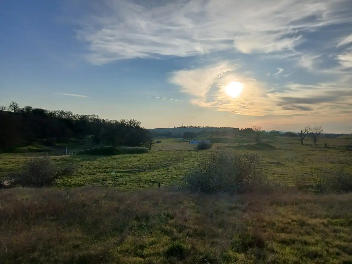 A scenic landscape photo of a grassy field with rolling hills, trees, and a bright sun partially obscured by wispy clouds in…