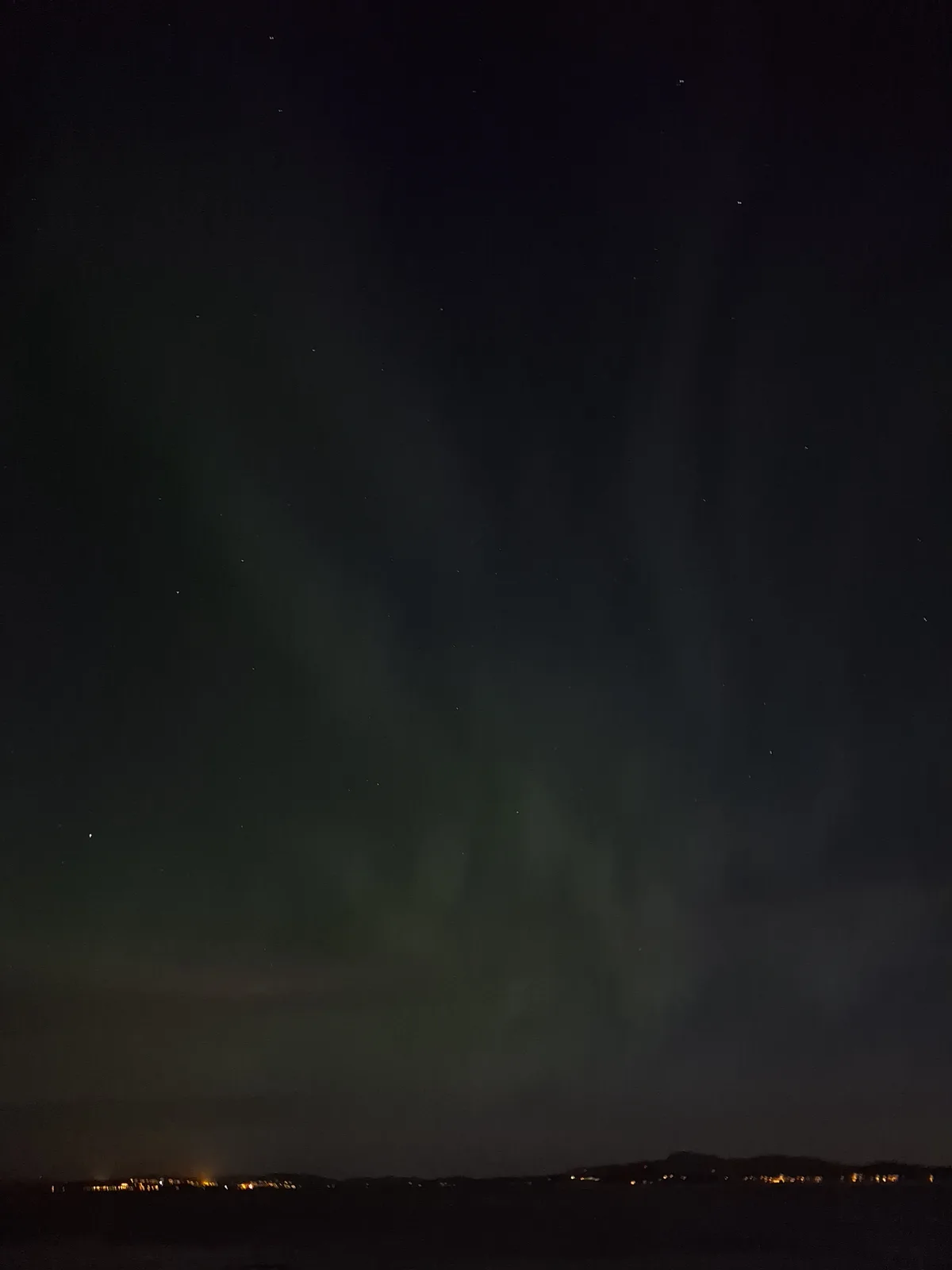 A night sky photograph showing faint green auroras dancing above a distant horizon dotted with city lights. The overlaid…