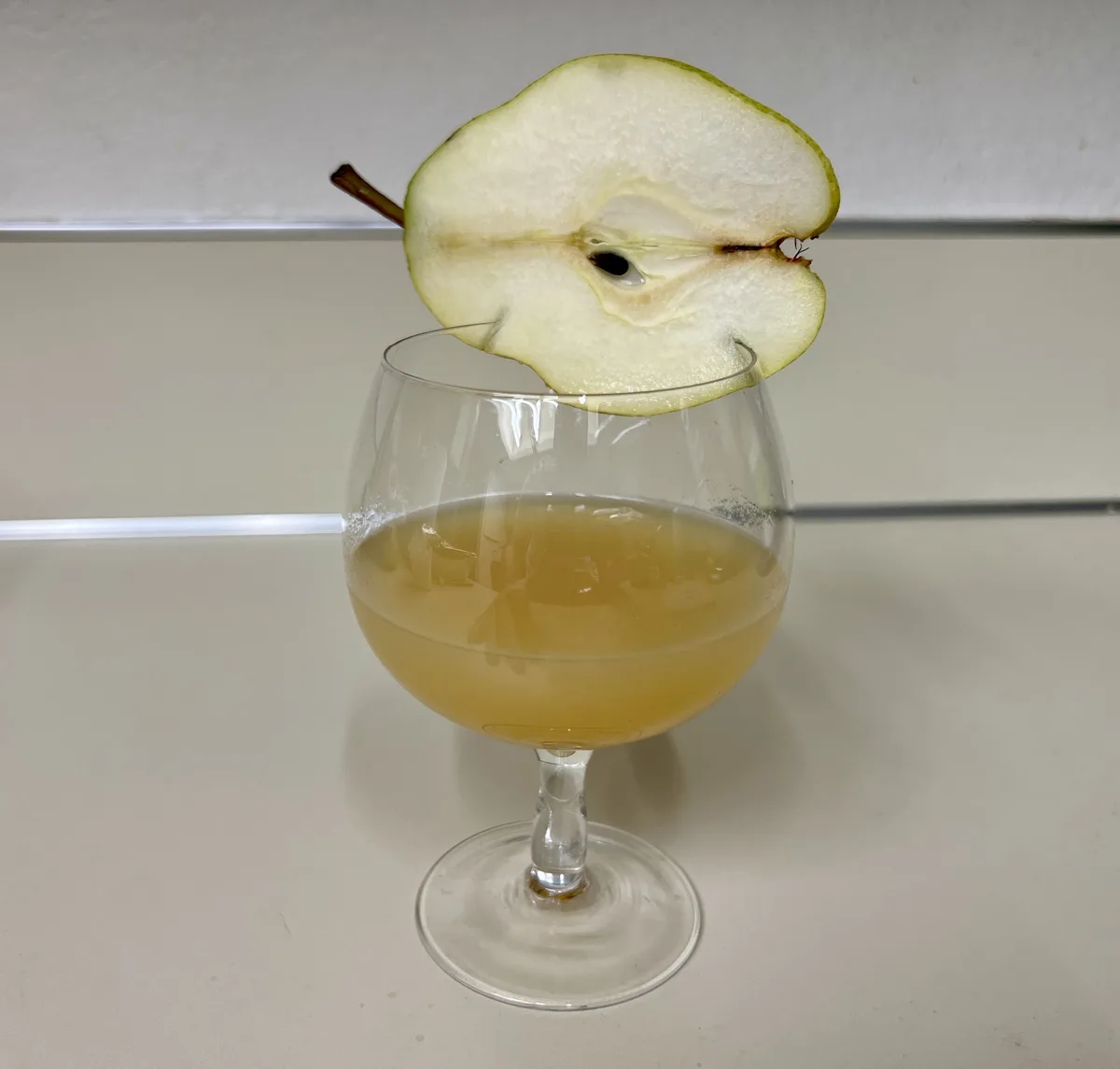 A clear glass filled with a pale yellow liquid sits on a light-colored countertop, garnished with a single slice of pear…