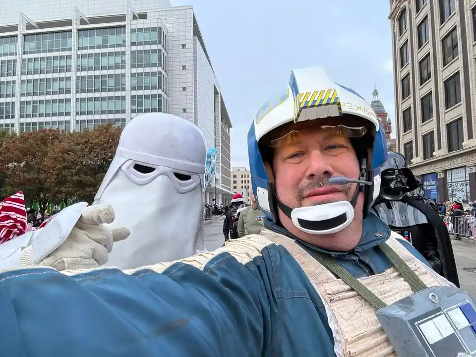 A man in a blue and white Star Wars pilot helmet and vest takes a selfie with a person in a white Stormtrooper costume. The…