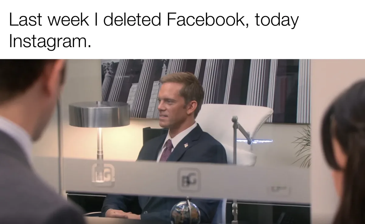 A man in a suit sits in an office chair behind a desk, looking slightly annoyed or exasperated while speaking to two people…