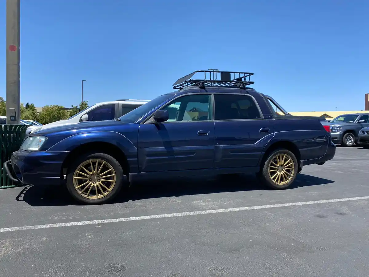 A dark blue Subaru Legacy wagon is parked in a sunny lot, featuring a black roof rack and distinctive gold-colored…