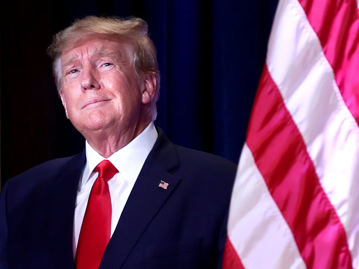 A man in a dark suit with a red tie and an american flag pin stands in front of a blue curtain and a partially visible…