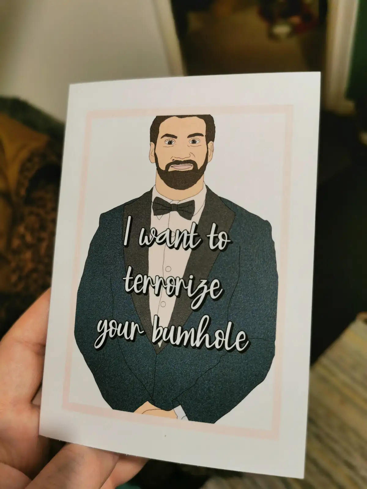 A hand holds a white card featuring a cartoon illustration of a man with a beard wearing a tuxedo and bow tie. Overlaid in…
