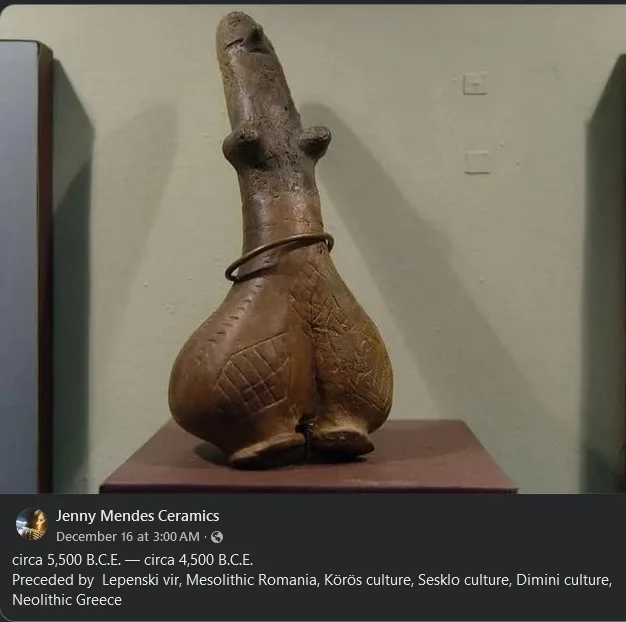 A brown, ancient ceramic figurine with a long, phallic protrusion and a ring around its midsection, displayed on a pedestal…