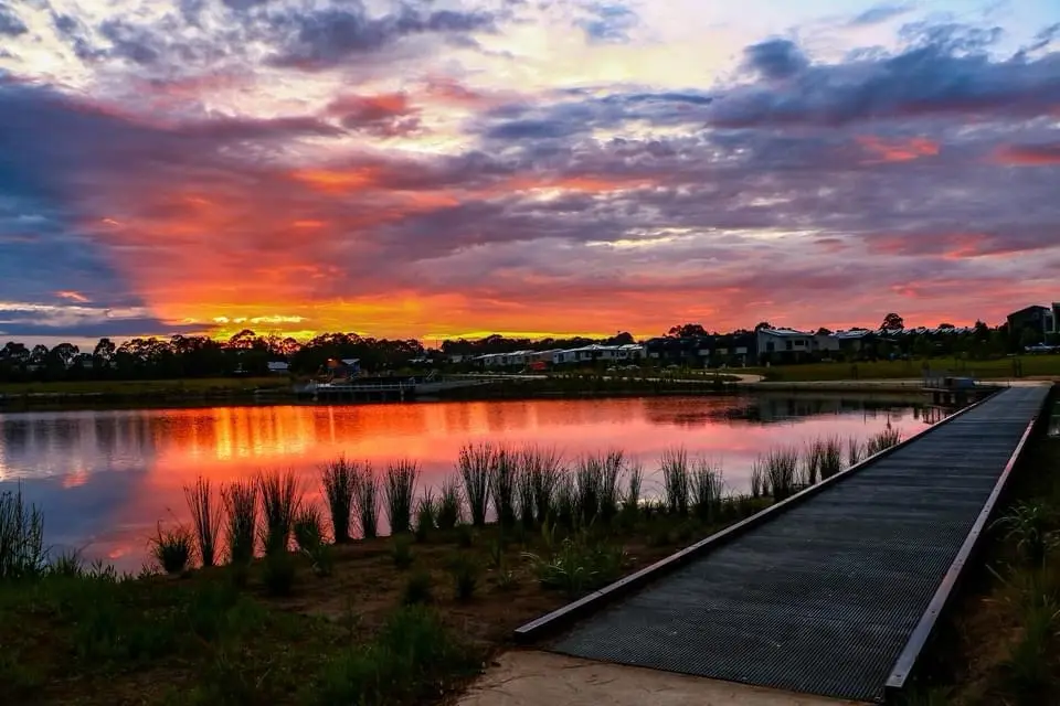 A serene sunset over a lake with a wooden boardwalk leading toward the water, reflecting vibrant orange and pink clouds…