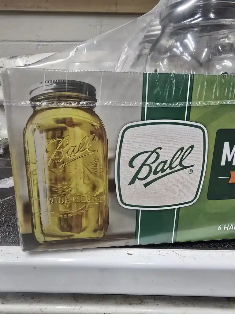 A package of Ball brand wide-mouth mason jars, with one jar visibly filled with a golden liquid, sits on a shelf. The…