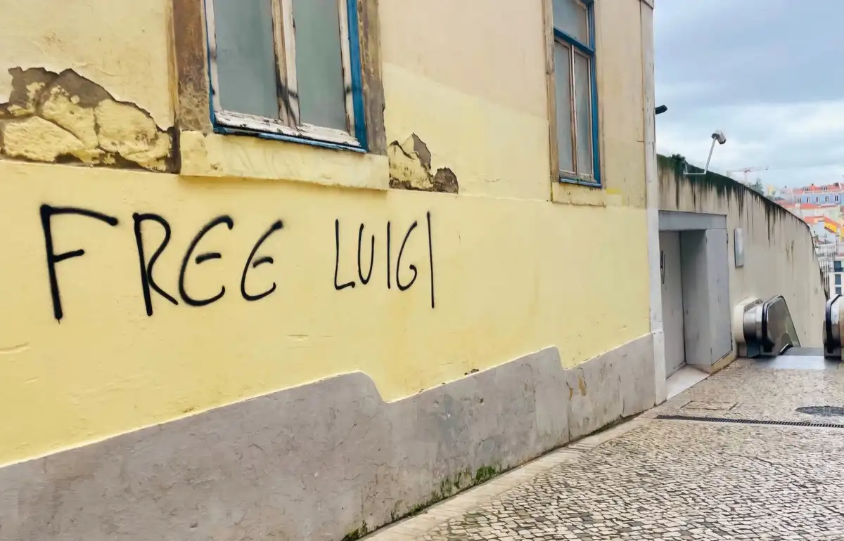 A yellow-painted wall on a cobblestone street has black graffiti reading 'FREE LUIGI'. The wall shows signs of wear and…