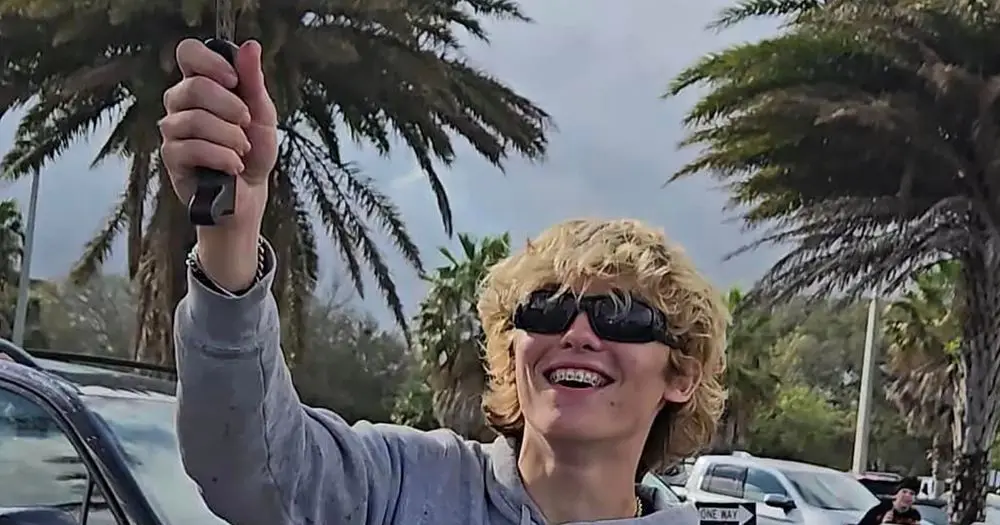 A person with curly blonde hair and sunglasses, smiling widely with braces, holds up a small black object in their right…