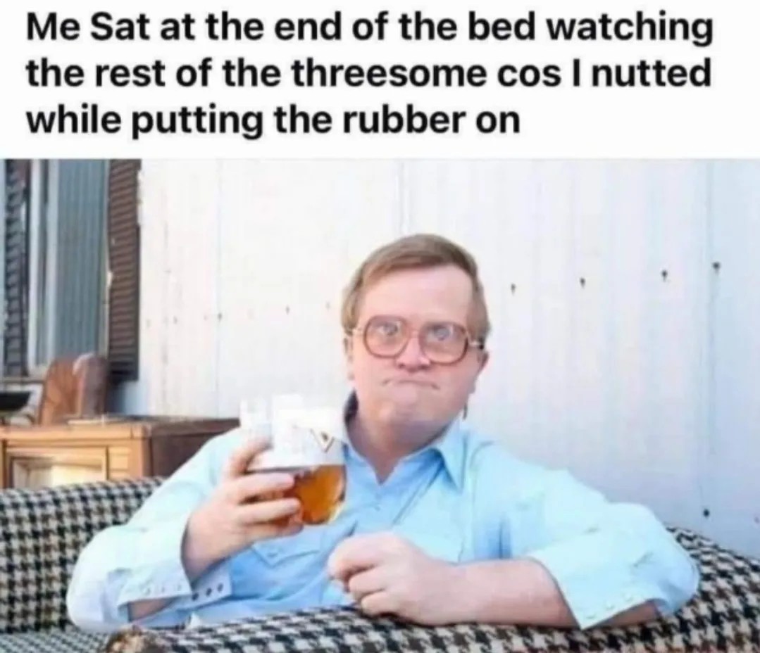 A man with glasses and a light blue shirt sits on a checkered couch holding a glass of amber liquid, looking smugly at the…