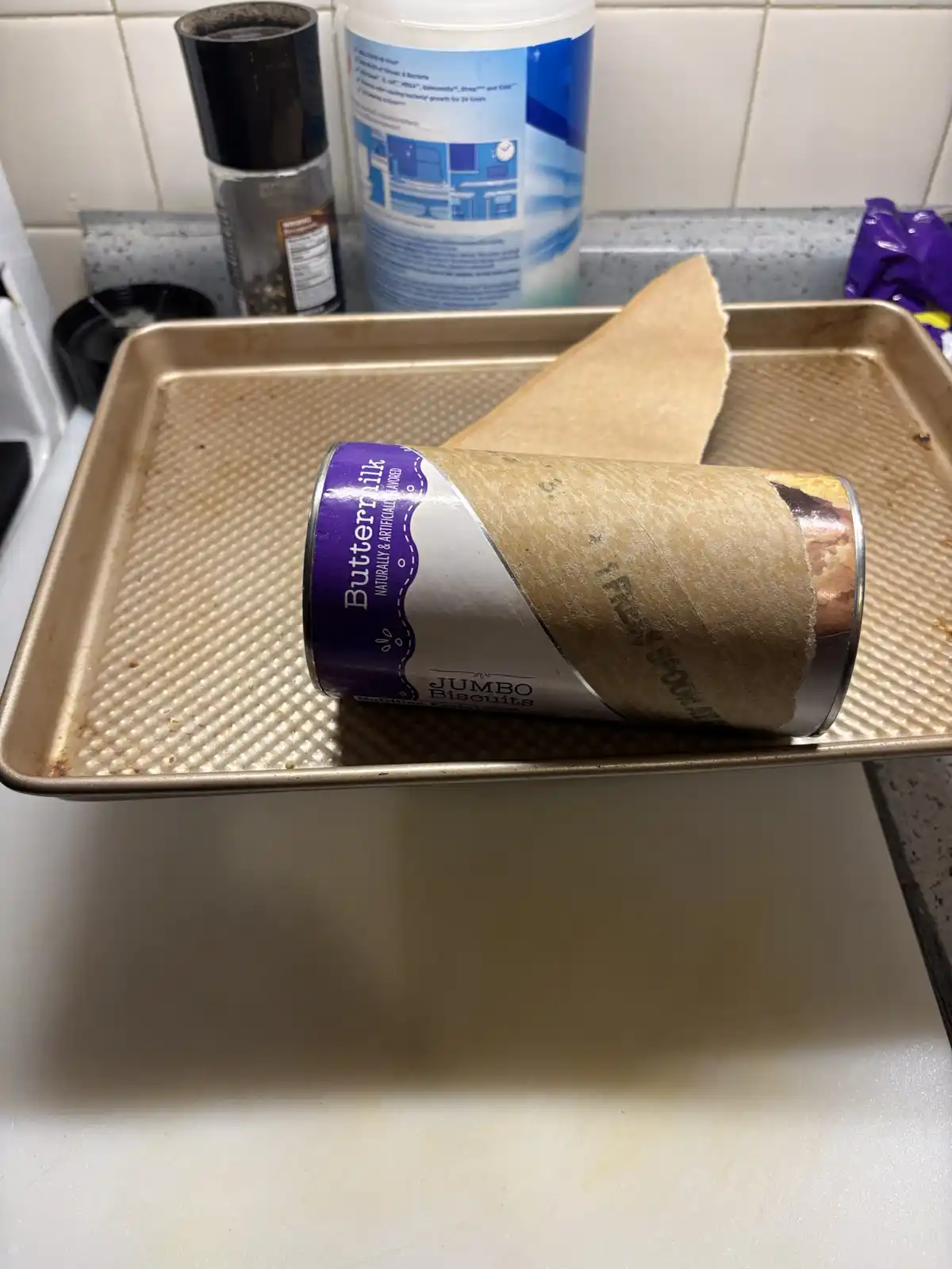 A can of Buttermilk Jumbo Biscuits lies on its side on a metal baking sheet, partially wrapped in brown paper. The can's…