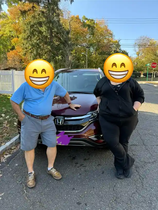 Two people with emoji faces standing proudly in front of a maroon Honda SUV on a sunny day with autumn trees in the…