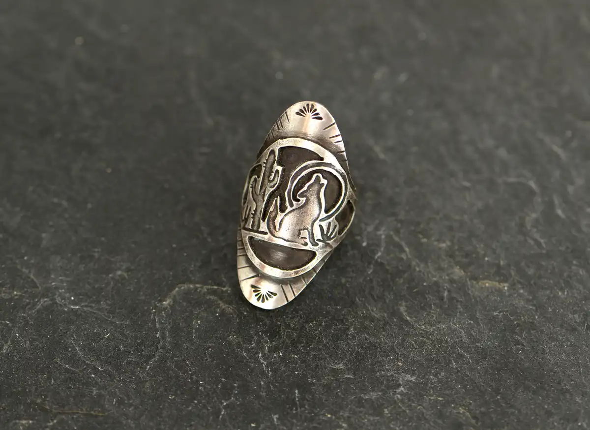 A silver ring with a carved design of a wolf howling at the moon, surrounded by cacti, rests on a dark slate surface. The…