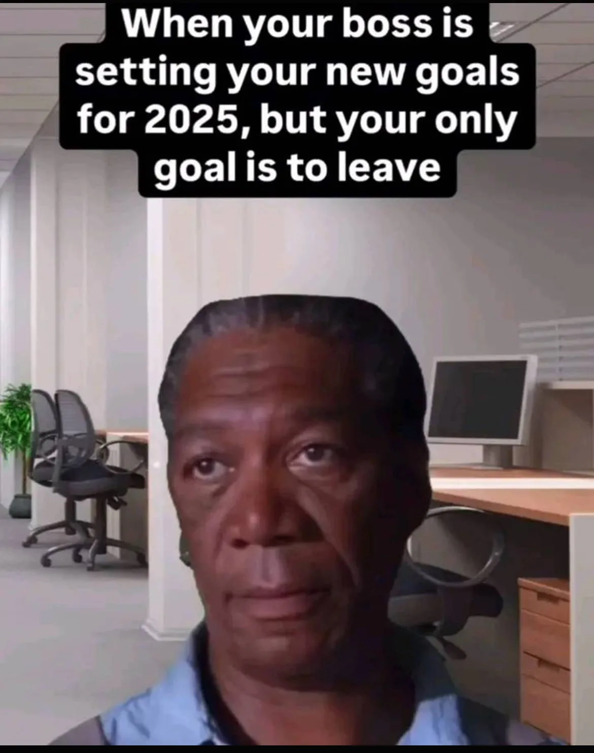 A man with a weary, resigned expression stares forward in an office setting with desks, chairs, and a computer monitor…