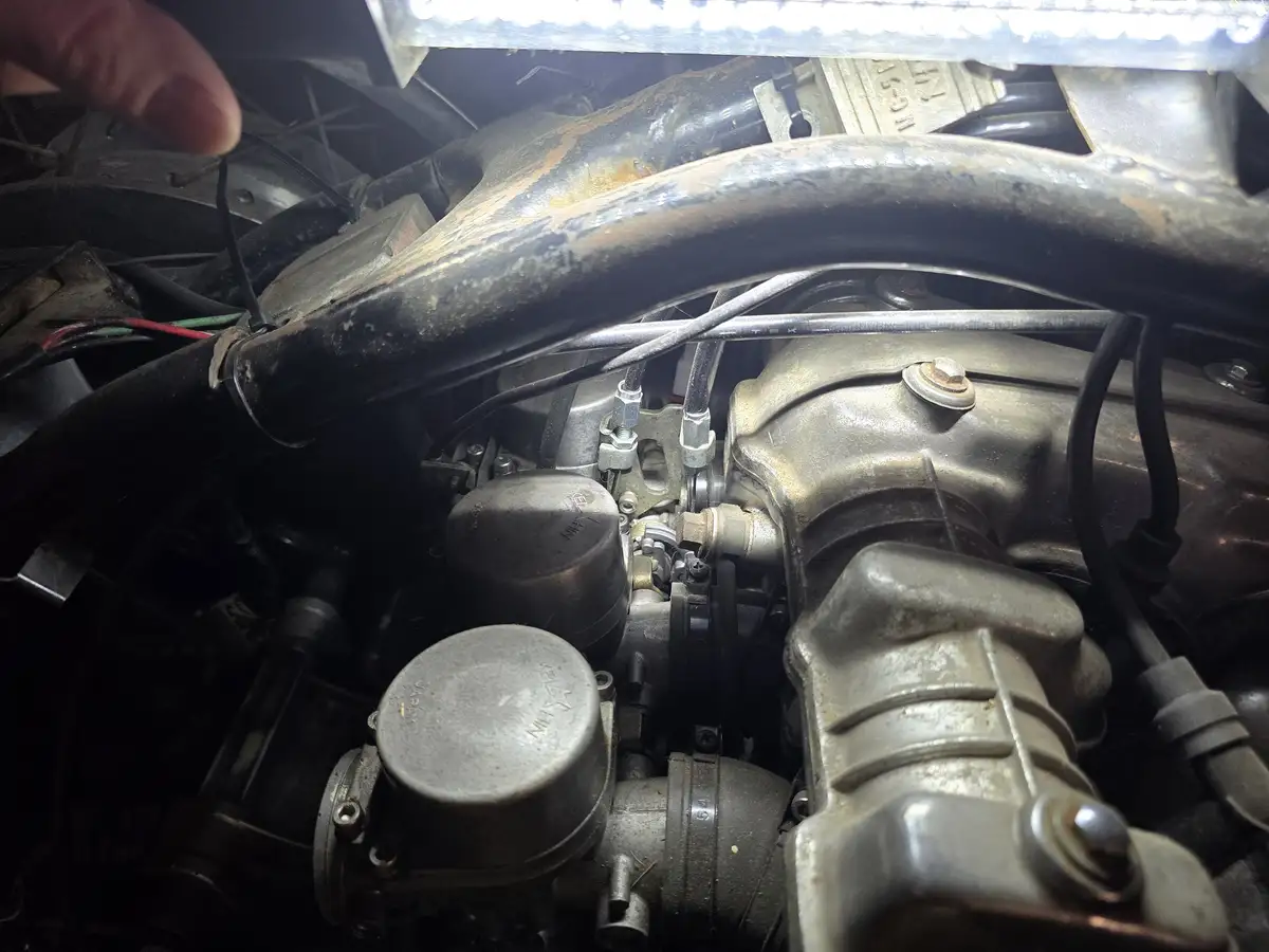 A close-up photo of a dirty, greasy car engine bay with a person's finger pointing at a component. The overlaid text reads…