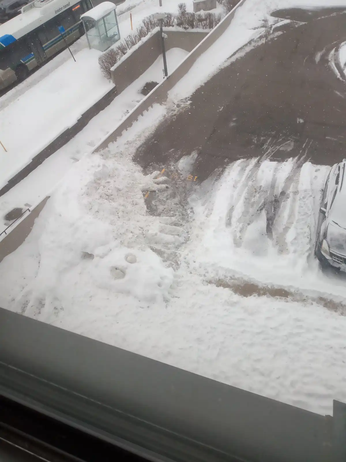 An overhead view from a window shows a parking lot covered in snow, with a snow-covered car on the right and a snow pile…