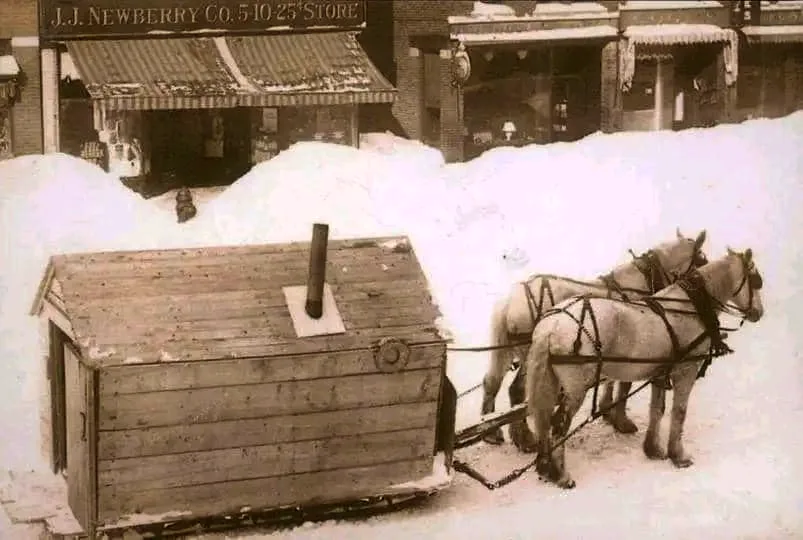 A sepia-toned historical photo shows two horses harnessed to a wooden sled shaped like a small house with a chimney, pulled…