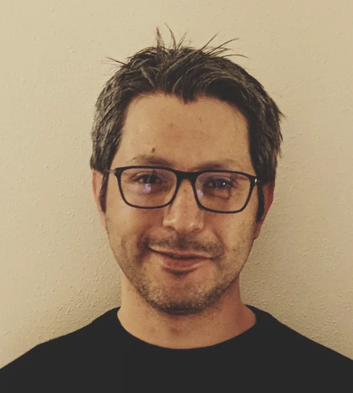 A man with dark hair, glasses, and a slight beard smiles faintly while wearing a black t-shirt against a plain beige wall…
