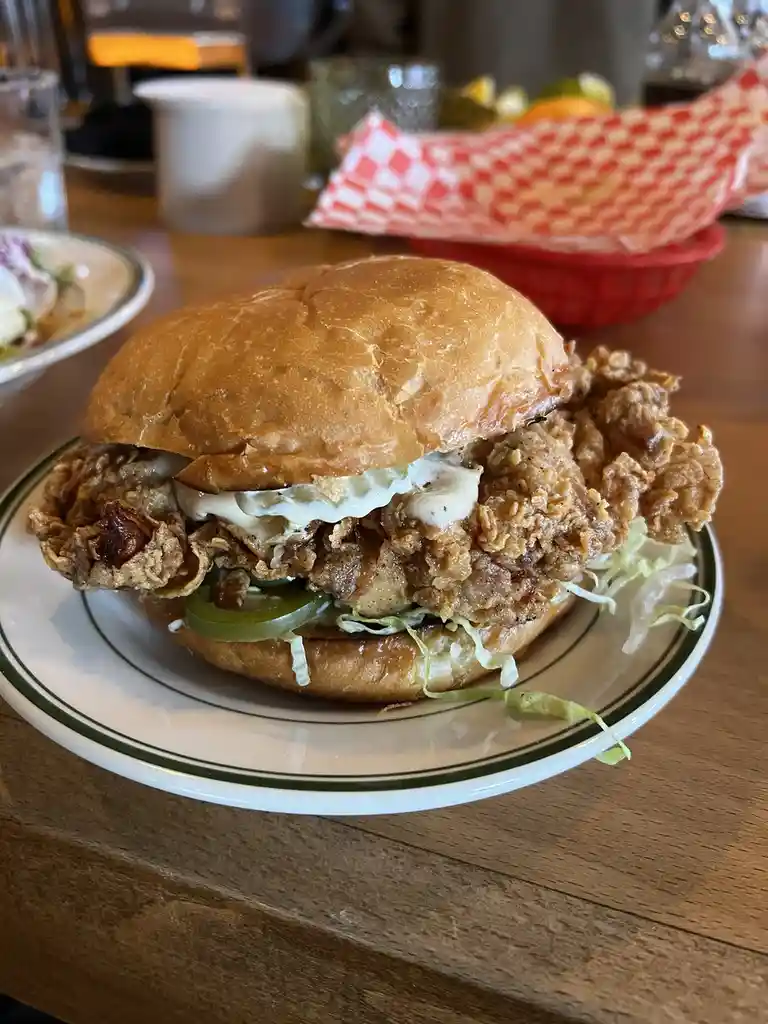 A close-up photo shows a large, crispy fried chicken sandwich on a soft bun, layered with lettuce, a green pepper slice, and…