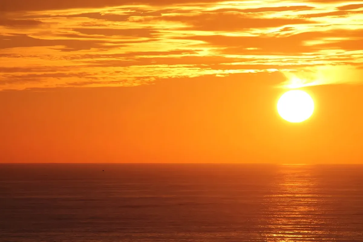A serene sunset over the ocean with the sun low on the horizon, casting orange and yellow hues across the sky and water. The…