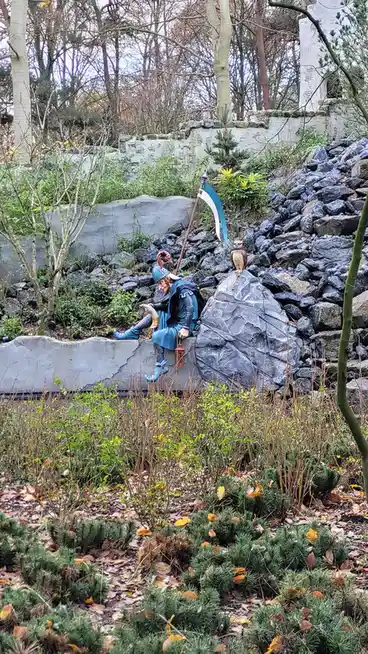 A statue of a blue-clad fantasy warrior sits on a stone ledge in a garden, holding a horn and a lantern, with a scythe and…
