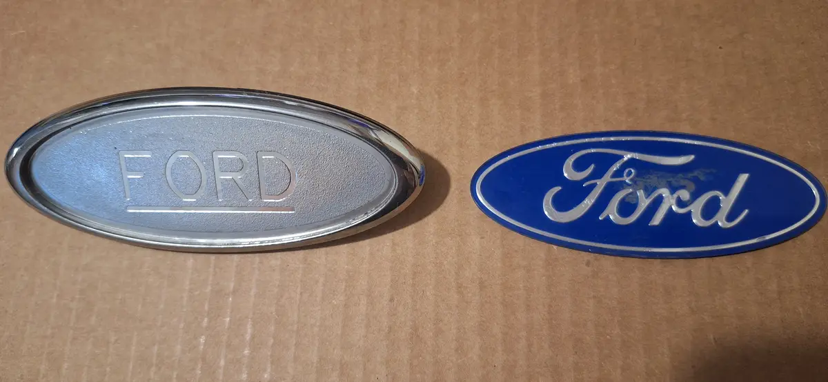 Two Ford car emblems lie side by side on a brown cardboard surface: one is a silver oval with 'FORD' in block letters, and…