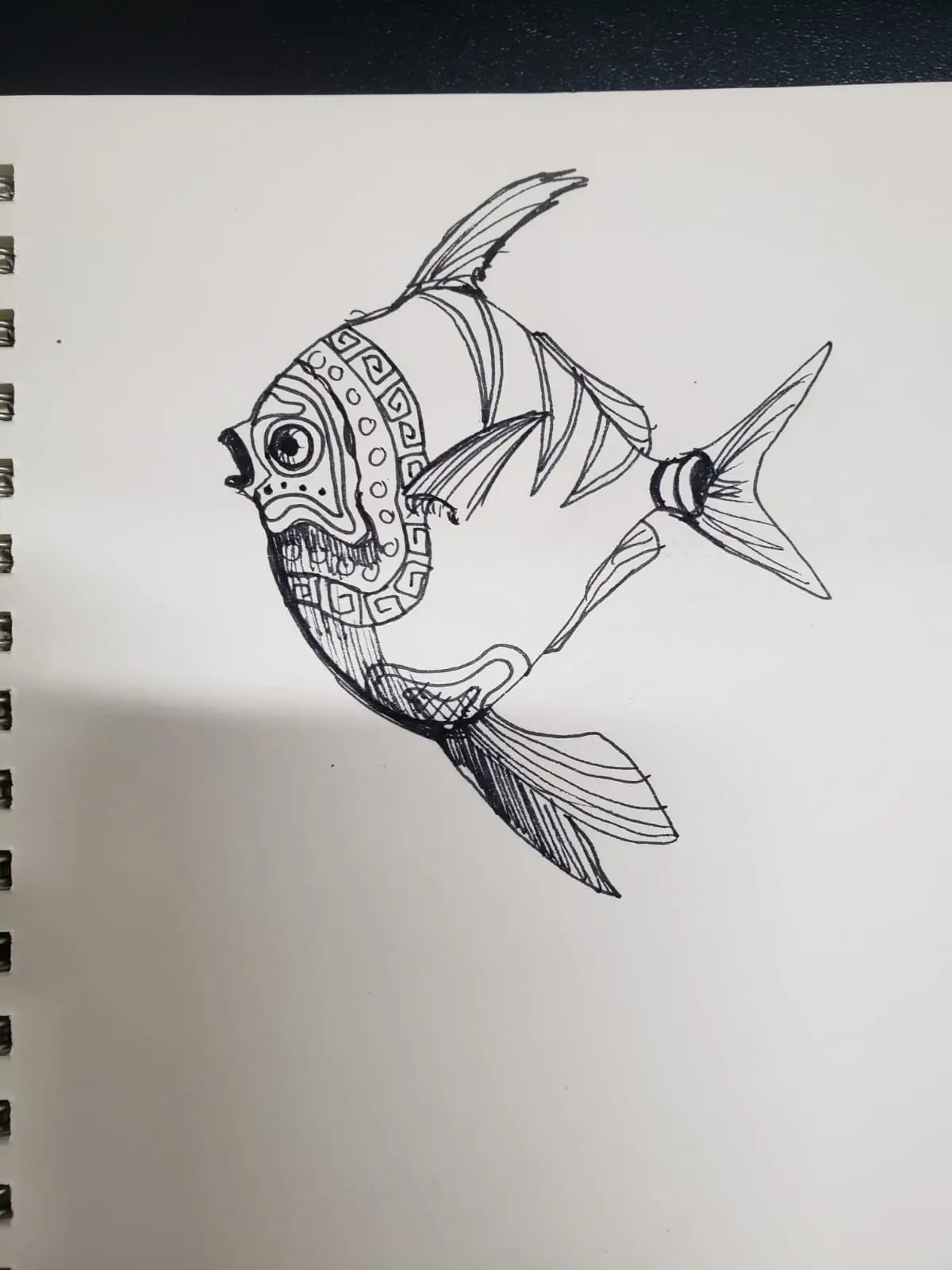 A black-and-white hand-drawn sketch of a stylized fish on a spiral-bound notebook page, featuring intricate geometric…