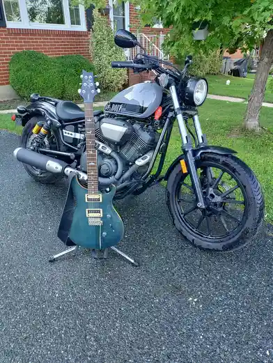 A dark gray motorcycle is parked on a wet asphalt driveway in front of a brick house, with a teal electric guitar leaning…