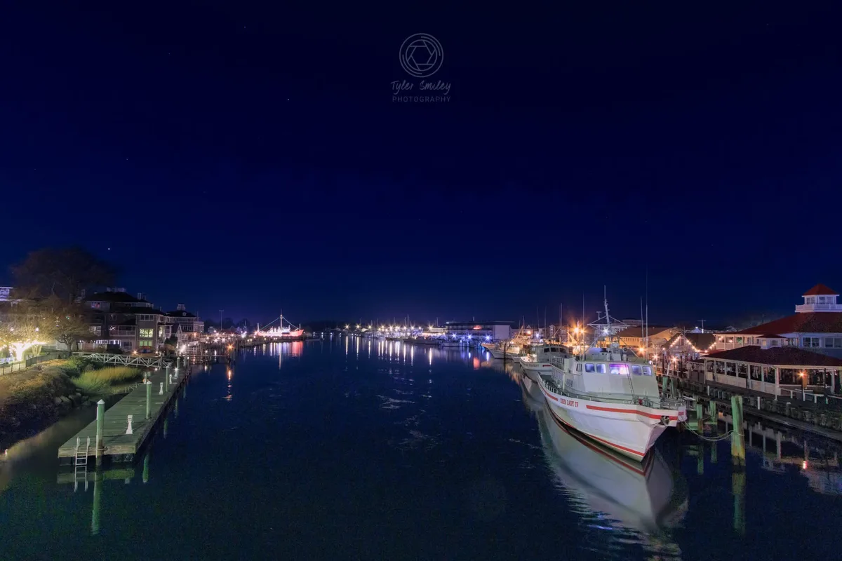 A serene night photograph of a harbor with boats docked along wooden piers, their lights reflecting on calm water under a…