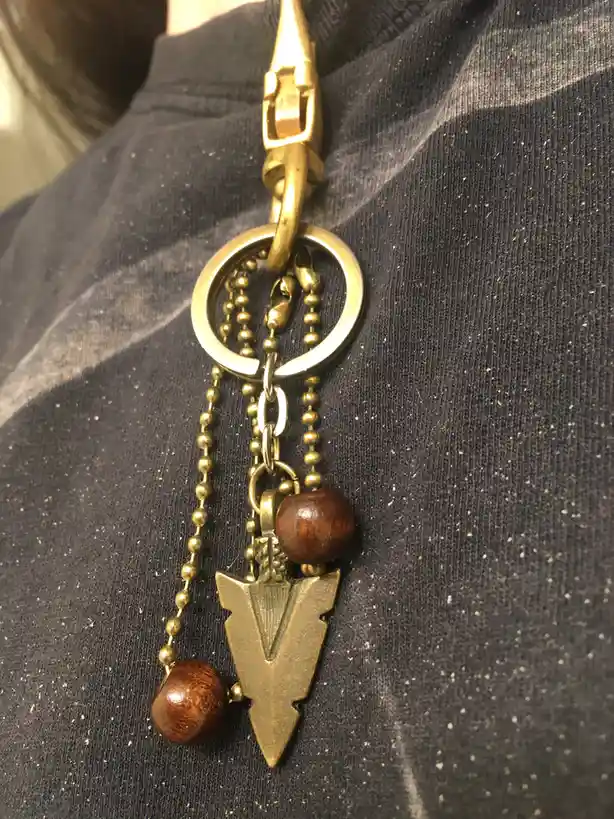 A close-up photo shows a brass-colored keychain with a large arrowhead charm, wooden beads, and ball chains, clipped to a…