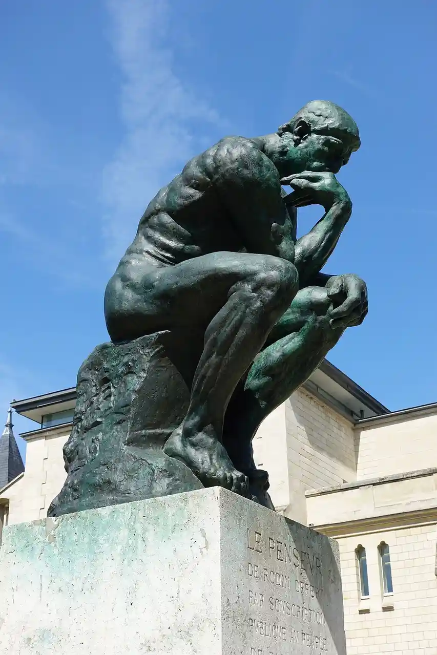 A bronze statue of a muscular, nude man in a crouched position with his chin resting on his hand, set against a blue sky…