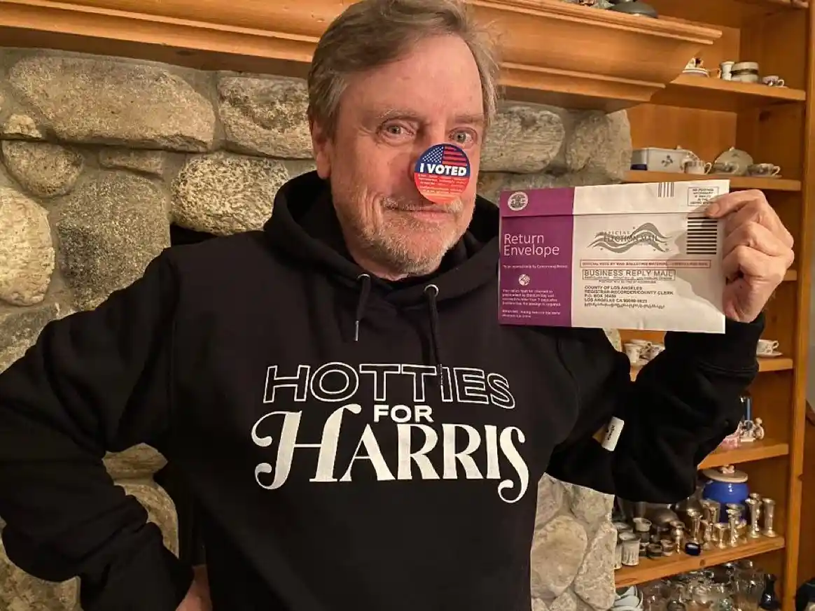 A man with gray hair and a goatee wears a black hoodie that reads 'HOTTIES FOR HARRIS' and holds up a mail-in ballot return…