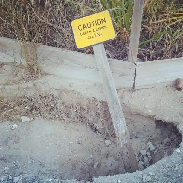 A yellow caution sign warning of 'BEACH EROSION CLIFFING' is visibly leaning at a severe angle, partially buried in sand and…