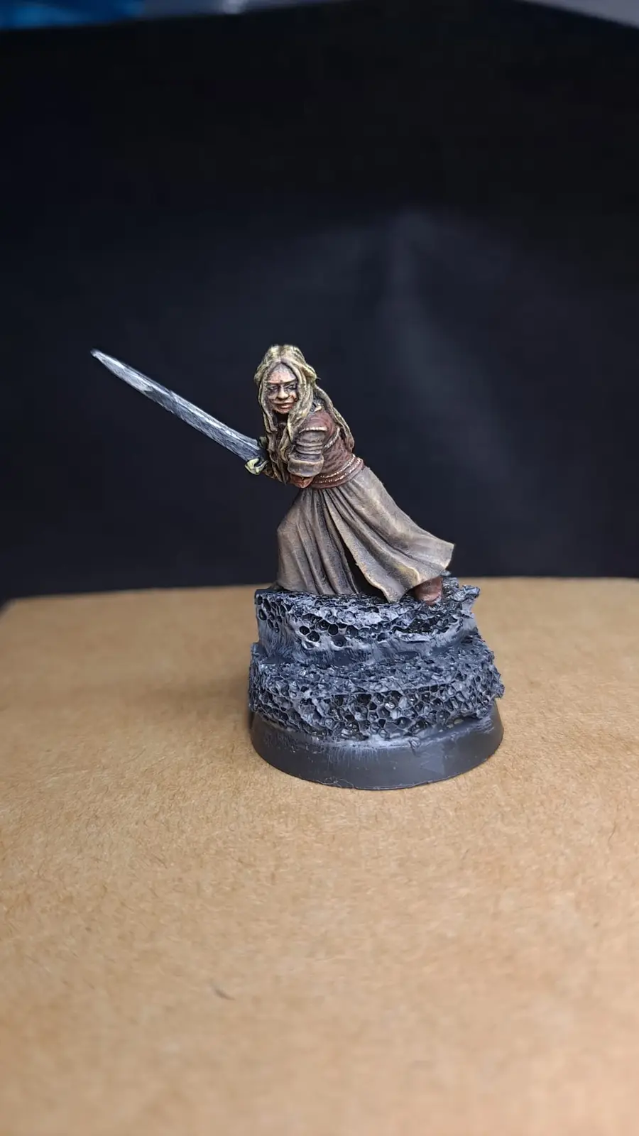 A painted miniature figurine of a female warrior with long blonde hair, wearing a brown tunic and skirt, standing on a rocky…